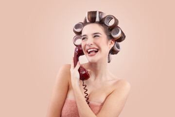 Happy woman with hair curlers, using a vintage telephone while look from the side. Concept of hairstyle, makeup, beauty routine, and cosmetics. Possibly Making a hair appointment. Isolated on beige