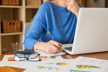 Businesswoman wearing a blue shirt is working from home, analyzing financial charts, taking notes, and using a laptop, with a calculator and eyeglasses on the desk