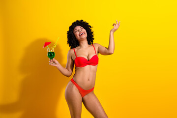 Positive young woman in vibrant red bikini enjoying a colorful cocktail against a cheerful yellow...