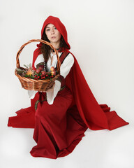 portrait pretty young female brunette model wearing a red hooded fairytale cloak and medieval historical gown. kneeling pose holding wicker basket. Isolated figure, white studio background