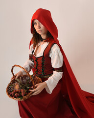 portrait pretty young female brunette model wearing a red hooded fairytale cloak and medieval historical gown. kneeling pose holding wicker basket. Isolated figure, white studio background