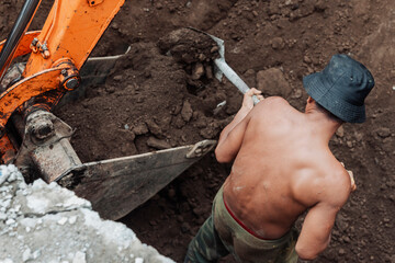 Shirtless construction worker using a shovel to move dirt into a excavator bucket in a trench...
