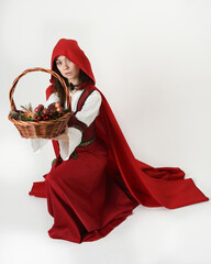 portrait pretty young female brunette model wearing a red hooded fairytale cloak and medieval historical gown. kneeling pose holding wicker basket. Isolated figure, white studio background