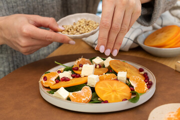 A woman pours pine nuts on a winter salad with persimmon, spinach and cheese. Healthy winter salad