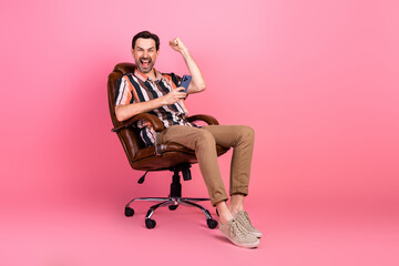 Fototapeta premium Cheerful man seated in chair celebrating success with phone against vibrant pink background in casual fashion