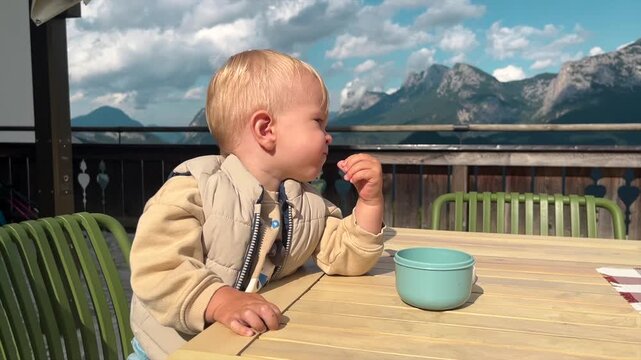 Little boy sits on a cafe terrace with mountain view and eats, he chews and frowns discontentedly from the sun. Children 1-2 years old, video with babies and toddlers. solid food introduction