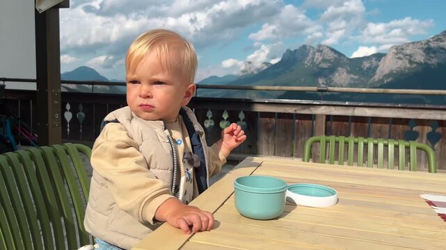 Little boy sits on a cafe terrace with Alps mountain view and eats, he chews and frowns discontentedly from the sun. Children 1-2 years old, video with babies and toddlers. solid food introduction