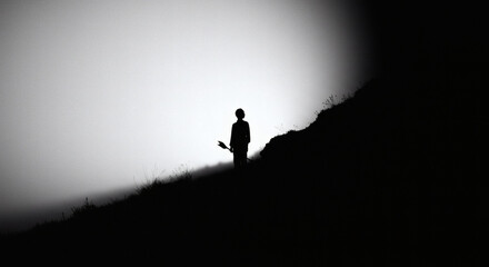 Fototapeta premium Silhouette of a person standing on a hillside, holding an object, against a foggy sky.