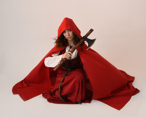 full length portrait  pretty young female brunette model wearing a red hooded fairytale cloak and medieval historical gown.  kneeling pose holding a axe weapon. Isolated figure, dark studio background