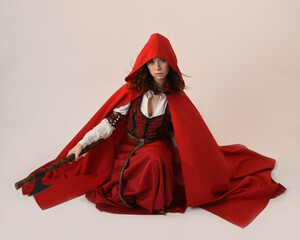 full length portrait  pretty young female brunette model wearing a red hooded fairytale cloak and medieval historical gown.  kneeling pose holding a axe weapon. Isolated figure, dark studio background