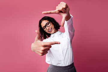 Smiling young woman in stylish white shirt making framing gesture with hands against pink backdrop...
