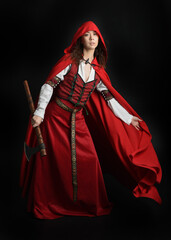 dramatic portrait of pretty young female brunette  model wearing a red hooded fairytale cloak and medieval historical gown. Holding a axe weapon. Isolated figure on dark studio background.