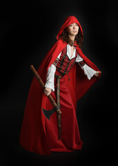 dramatic portrait of pretty young female brunette  model wearing a red hooded fairytale cloak and medieval historical gown. Holding a axe weapon. Isolated figure on dark studio background.