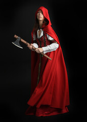 dramatic portrait of pretty young female brunette  model wearing a red hooded fairytale cloak and medieval historical gown. Holding a axe weapon. Isolated figure on dark studio background.