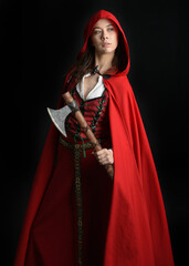 dramatic portrait of pretty young female brunette  model wearing a red hooded fairytale cloak and medieval historical gown. Holding a axe weapon. Isolated figure on dark studio background.