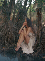 Young woman in a white dress sits barefoot by a forest lake, among tree roots and reflected in the...
