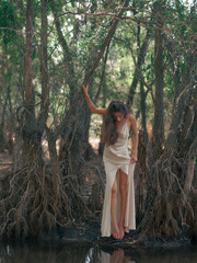 Young woman in a white dress stands barefoot by a forest lake, among tree roots and reflected in...