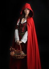 dramatic portrait of pretty young female brunette  model wearing a red hooded fairytale cloak and medieval historical gown. Holding a wicker basket. Isolated figure on dark studio background.