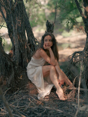 Young woman in white dress sits barefoot in mysterious forest, surrounded by old tree roots.