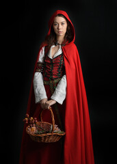 dramatic portrait of pretty young female brunette  model wearing a red hooded fairytale cloak and medieval historical gown. Holding a wicker basket. Isolated figure on dark studio background.