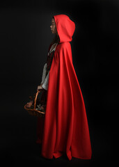 dramatic portrait of pretty young female brunette  model wearing a red hooded fairytale cloak and medieval historical gown. Holding a wicker basket. Isolated figure on dark studio background.