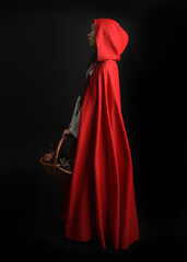 dramatic portrait of pretty young female brunette  model wearing a red hooded fairytale cloak and medieval historical gown. Holding a wicker basket. Isolated figure on dark studio background.