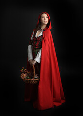 dramatic portrait of pretty young female brunette  model wearing a red hooded fairytale cloak and medieval historical gown. Holding a wicker basket. Isolated figure on dark studio background.