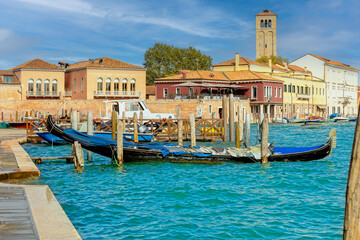 Traditional Venetian waterfront scene on Murano island with gondolas, colorful buildings, and a historic bell tower under sunny skies.