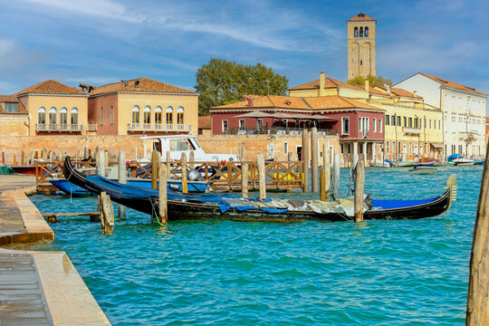 Traditional Venetian waterfront scene on Murano island with gondolas, colorful buildings, and a historic bell tower under sunny skies.
