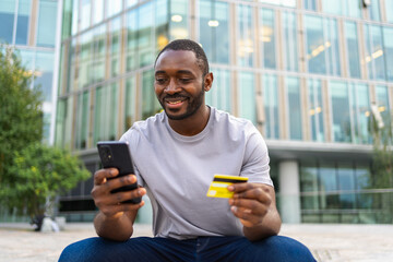 African American man shopping online holding smartphone paying with gold credit card Guy on urban street in city buying on Internet enter credit card details Online shopping ecommerce delivery service