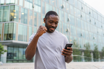 Happy African American man euphoric winner with smartphone on street in city. Person guy looking at cell phone reading great news getting good result winning online bid feeling amazed. Winning gesture