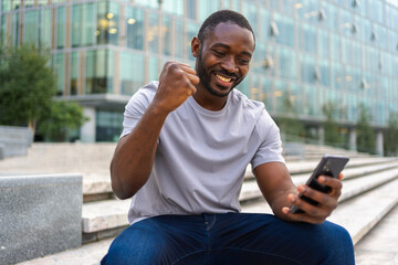 Happy African American man euphoric winner with smartphone on street in city. Person guy looking at...