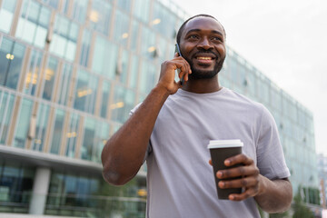 Good morning. African man drinking coffee to go talking on smartphone outdoor. Man with cell phone paper cup walking on street. Man making answering call by cellphone having conversation by mobile