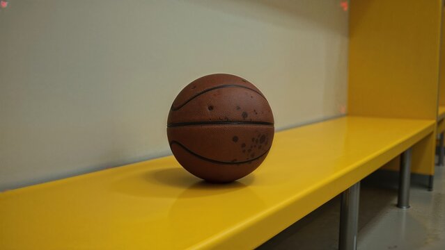A brown basketball positioned on a yellow bench within a locker room setting. Horizontal sports graphic. - Powered by Adobe