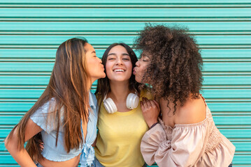 Two multiracial young women kissing the cheeks of their friend against a blue background