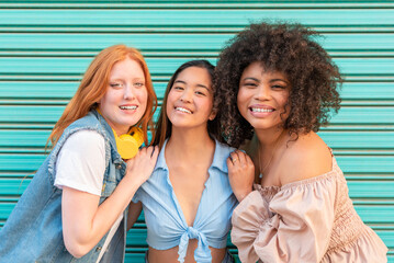 Three cheerful multi-ethnic female friends embracing against a turquoise wall smiling at camera