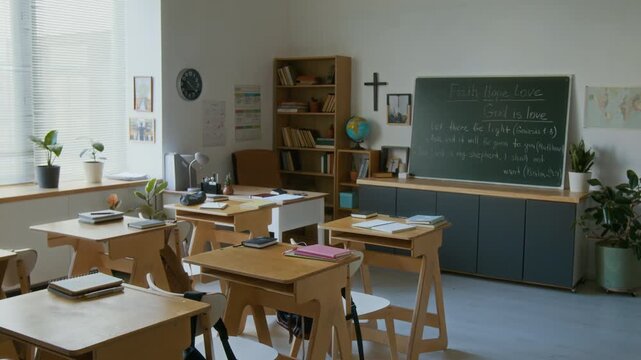 No people shot of empty classroom with single school desks and chairs in modern religious school with Cristian cross on wall and Bible quotes on blackboard