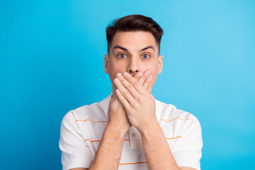 Young man with a surprised expression covering mouth with hands against a vibrant blue background...