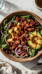 Overhead view of a vibrant salad with avocado bacon red onion and greens in a wooden bowl on a table