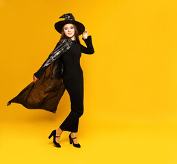Cheerful young woman witch wearing black dress, hat, cloak on yellow background. Concept halloween...