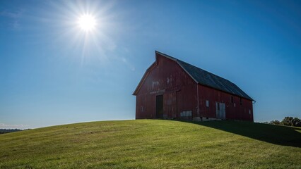Obraz premium On a verdant hill, an old red barn with a slanting roof basks in the sunlight under a clear azure sky.
