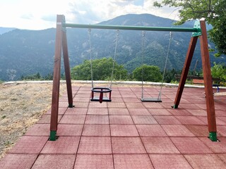 Two empty swings on red playground tiles, mountains and green trees in the scenic background