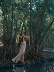 Young woman in a white dress stands barefoot by a forest lake, among tree roots and reflected in...