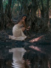 Young woman in white dress sits and rests by forest lake among old tree roots.