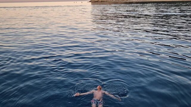 Relaxed person floats on his back in clear blue water at sunset. Serene Kemer coastal view with majestic Taurus mountains in the background. Summer holiday chill as man swims in the Mediterranean sea