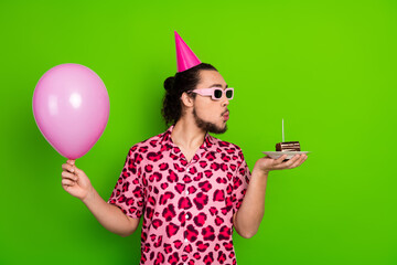 Stylish young man celebrating with pink balloon and party cake on vibrant green backdrop,...