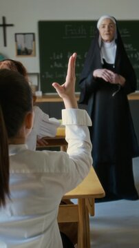Vertical rear view shot of student girl sitting at desk in classroom raising hand during lesson with mature Caucasian nun in veil teaching in modern Christian school