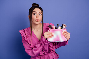 Charming young woman in vibrant pink dress holding striped makeup pouch against vibrant violet...