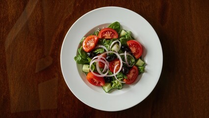 Fresh vegetable medley with tomatoes, cucumber, onions, leafy salad greens, and black olives served on a white plate over a dark wooden table.