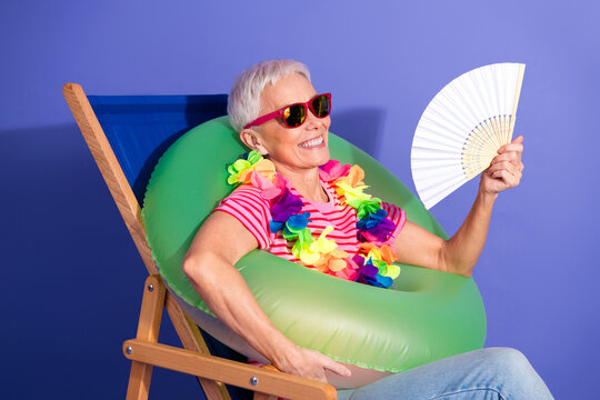Cheerful senior woman in colorful summer attire relaxing with a green float and fan against a purple background - Powered by Adobe